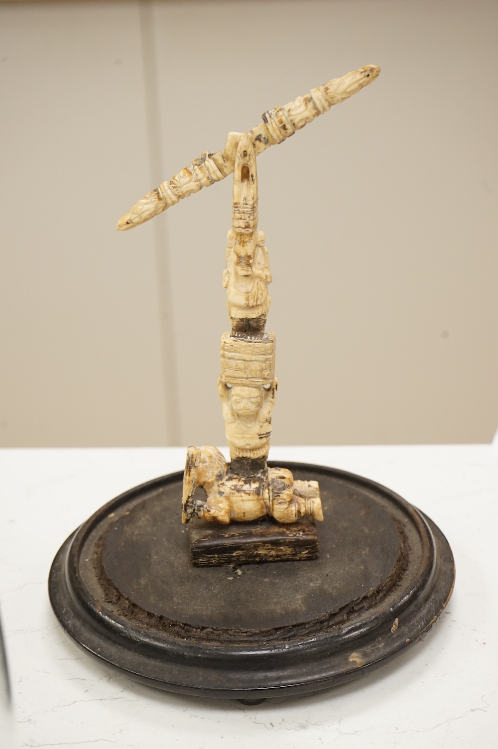 A 19th century Indian bone sculptural devotional object, carved as a totem with Ganesh and other deities, pivoting beam, 18cm high, glass dome with ebonised base